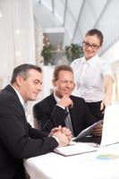 It was a good deal. Two cheerful men in formalwear sitting at the table while woman in white shirt standing close to them and pointing computer monitor photo