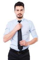 Everything should be perfect. Confident young man in shirt and tie looking at camera and adjusting his necktie while standing against white background photo