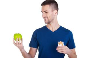 I made my choice. Confident young man looking at the apple in his hand while holding pack of cigarettes in another hand and isolated on white photo