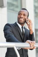 feliz de que el negocio vaya. apuesto joven africano en ropa formal hablando por teléfono móvil y sonriendo mientras está de pie al aire libre foto