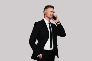 Glad to hear you Good looking young man in full suit talking on the phone and smiling while standing against grey background photo