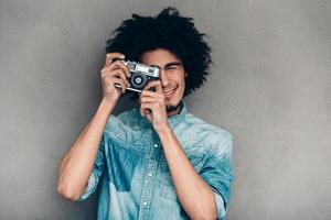 Show me your best smile Cheerful young African man holding retro styled camera and focusing on you while standing against grey background photo