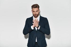 Confident in his style. Handsome young man in formalwear holding hands clasped and looking at camera while standing against grey background photo