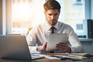 Checking his timetable. Pensive young handsome man using his digital tablet while sitting at his working place photo