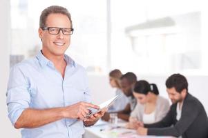 Confident team leader. Confident young man in glasses working on digital tablet and smiling while people working on background photo