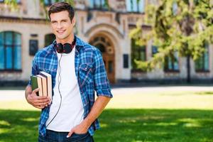 estudiante confiado. apuesto joven sosteniendo libros y sonriendo mientras está de pie frente a su universidad foto