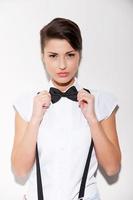 Confident and stylish. Beautiful young short hair woman in white shirt and suspenders adjusting her bow tie and looking at camera photo