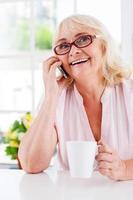 Good news. Cheerful senior woman talking on the mobile phone and smiling while sitting at the table photo