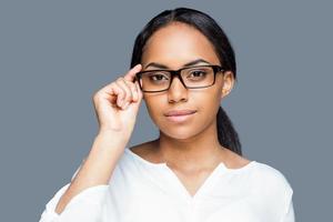 Confident look. Attractive young African woman adjusting her eyeglasses and looking at camera while standing against grey background photo