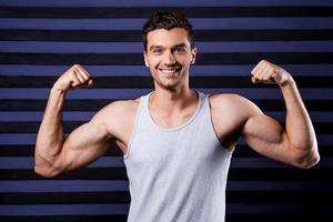 musculoso guapo. joven musculoso en la parte superior del tanque mostrando sus bíceps perfectos y sonriendo mientras está de pie contra el fondo rayado foto