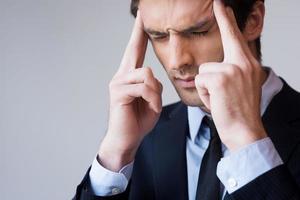 Stressed and overworked. Frustrated young man in formalwear touching head with fingers and keeping eyes closed while standing against grey background photo