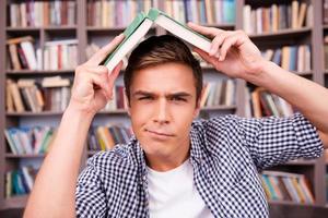 Tired of studying. Frustrated young man carrying book on head and expressing negativity while sitting against bookshelf photo