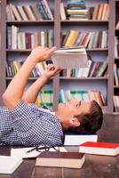 Happy bookworm. Side view of happy young man reading book while lying on the floor against bookshelf photo