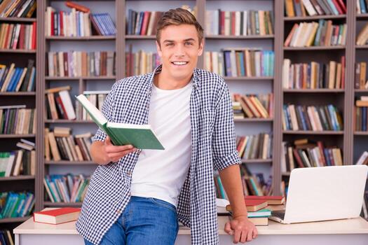 Smart and confident. Confident young man holding book and smiling while leaning at the desk in library photo