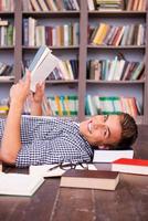 Happy bookworm. Side view of happy young man reading book while lying on the floor against bookshelf photo