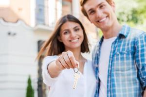 Moving to a new house. Beautiful young loving couple standing against house while woman holding key and smiling photo