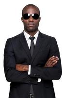 Cool and handsome. Serious young African man in formalwear and sunglasses keeping arms crossed and looking at camera while standing isolated on white background photo