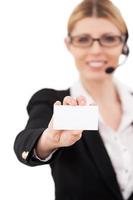 Copy space on her card. Confident mature customer service representative in headset stretching out hand with business card and smiling while standing isolated on white photo