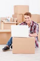 Searching a moving company in the net. Cheerful young man sitting on the floor and working on laptop while cardboard boxes laying in the background photo