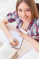 Smart and confident schoolgirl. Top view of cheerful teenage girl studying while sitting at the desk photo