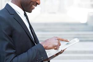 Working on tablet. Side view cropped image of African man in formalwear working on digital tablet and smiling while standing outdoors photo