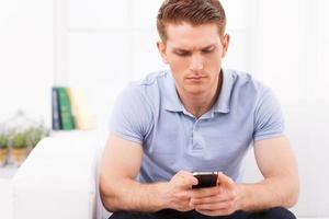 Bad news. Depressed young man holding mobile phone and looking at it while sitting on the couch photo