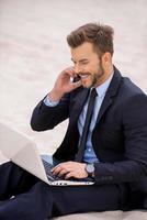 él siempre está en contacto. alegre joven hombre de negocios trabajando en una laptop y hablando por teléfono móvil mientras se sienta en la arena foto