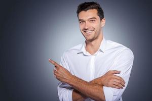 Handsome pointing copy space. Happy young man in white shirt looking at camera and smiling while standing against grey background and pointing copy space photo