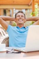 Dreaming of vacation. Relaxed mature man holding hands behind head and smiling while sitting at the table outdoors with laptop on it photo