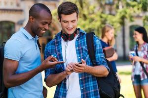 Sharing news with friends. Two cheerful men looking at the mobile phone and smiling with two women talking in the background photo