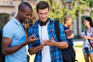 Sharing news with friends. Two cheerful men looking at the mobile phone and smiling with two women talking in the background photo