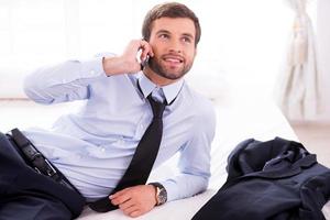 Executive on the go. Handsome young man in shirt and tie talking on the mobile phone and smiling while lying in bed photo