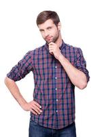 Thinking about solutions for you. Thoughtful young man in casual shirt holding hand on chin and looking at camera while standing isolated on white photo