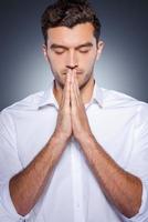 Praying for business. Handsome young man in white shirt holding hands clasped near face while standing against grey background photo