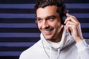 Music is my life. Handsome young man in white sweater adjusting his headphones and smiling while listening to the music and standing against striped background photo