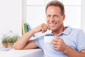 Relaxing with fresh coffee and latest news. Cheerful mature man drinking coffee and reading newspaper while sitting on the couch at home photo