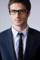 Portrait of success. Portrait of confident young man in formalwear and glasses looking at camera while standing against grey background photo