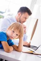 Feeling lonely. Depressed little boy holding hand in hair and looking at camera while his busy father working in the background photo