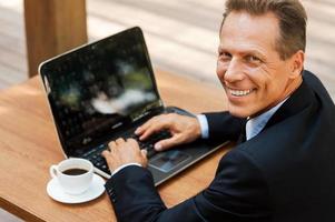 Me encanta trabajar al aire libre. vista superior de un hombre maduro alegre con ropa formal que trabaja en una laptop y sonríe mientras se sienta en un café al aire libre foto