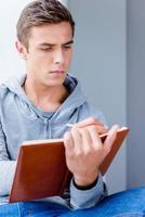 Young and creative. Thoughtful young man writing something in his note pad while sitting on the window sill photo