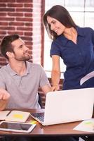 Helping out a colleague. Cheerful young man sitting at his working place and gesturing while beautiful woman standing close to him and smiling photo