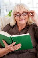 leyendo su libro favorito. anciana leyendo un libro y ajustándose las gafas mientras se sienta en la silla foto