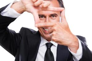 Businessman focusing at you. Playful mature man in formalwear gesturing finger frame and looking through it while standing isolated on white background photo