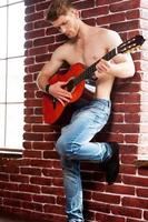 Playing guitar. Handsome young shirtless man playing acoustic guitar while leaning at the brick wall