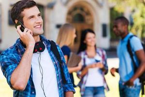 hablando con amigos. apuesto joven hablando por teléfono móvil y sonriendo mientras está de pie contra el edificio de la universidad con sus amigos charlando en el fondo foto