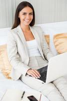 Staying in touch with office. Top view of confident young businesswoman in suit working on laptop and smiling while sitting in bed at the hotel room photo