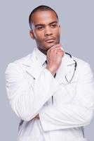 Pensive doctor. Thoughtful African doctor holding hand on chin and looking away while standing against grey background photo