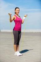 Ready to jump. Full length of beautiful young woman in sports clothing holding jumping rope and smiling while standing outdoors photo