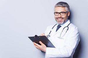 Writing prescription. Cheerful mature doctor looking at camera and smiling while writing something at his clipboard and standing against grey background photo