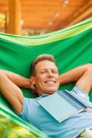 So good to do nothing. Happy mature man sleeping while lying in hammock with book laying on his torso photo